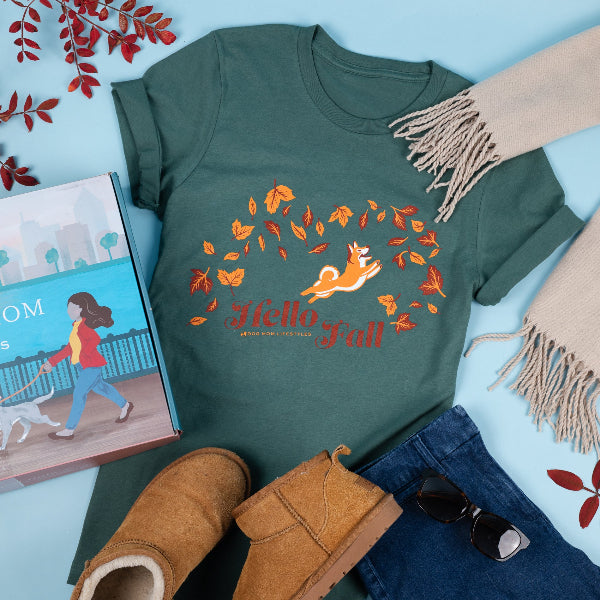 Green t-shirt with 'Hello Fall' design featuring a fox and leaves, surrounded by autumn-themed items on a light blue background.