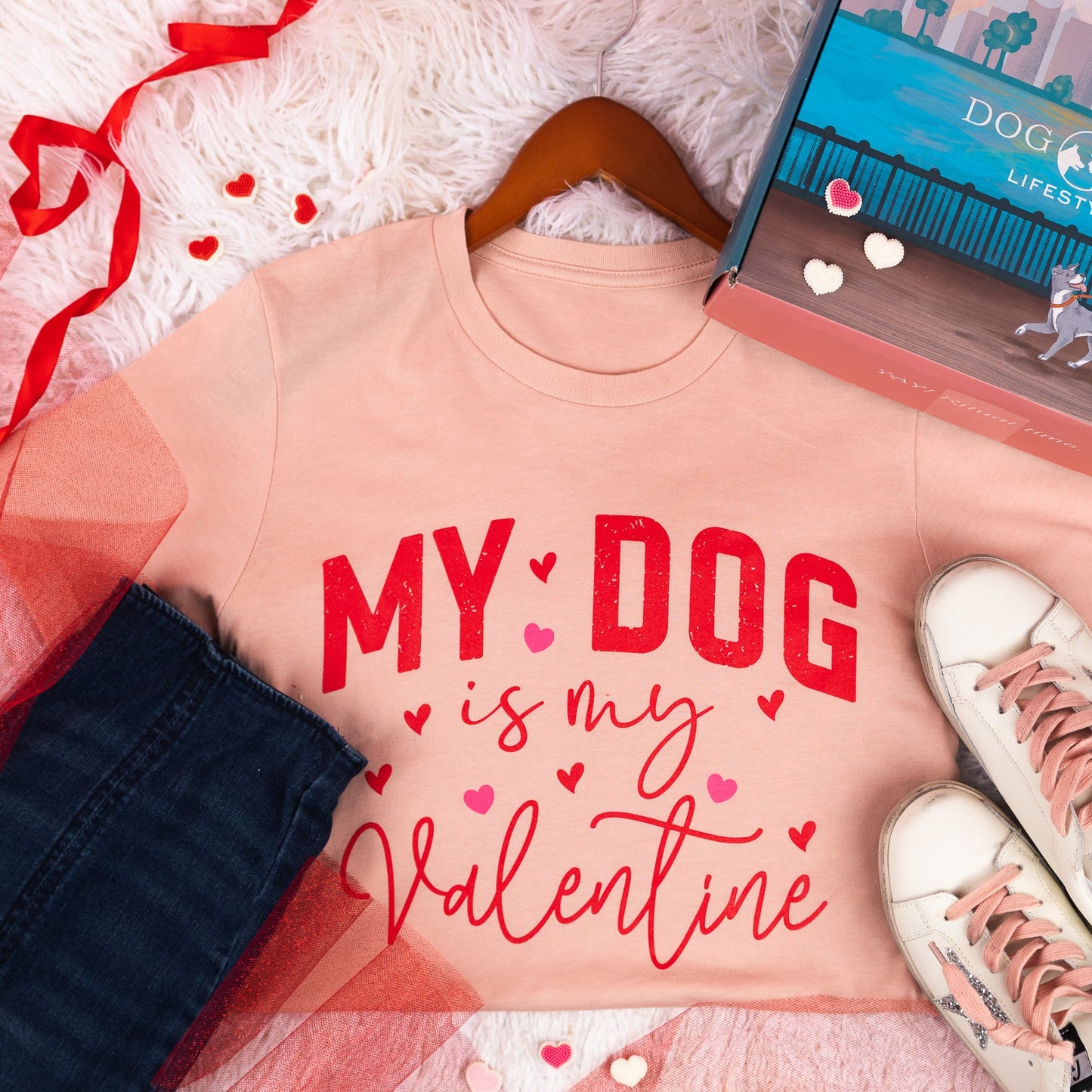 Pink t-shirt with 'My Dog is my Valentine' text, white sneakers, and a box labeled 'Dog Mom Lifestyles' on a textured surface.
